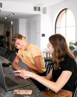 two women with smiling faces sitting side by side talking in front of their laptop