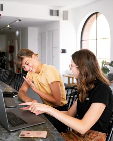 two women with smiling faces sitting side by side talking in front of their laptop